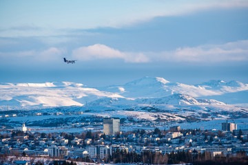 The plane is landing in a Reykjavik downtown in a golden evening light. Capital city of Iceland Reykjavik in winter surrounded with snow covered mountains.