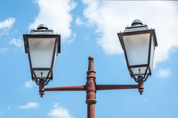 Fototapeta na wymiar Old vintage and rusty street lamp post or lantern with two light bulbs against beautiful blue sky with white clouds background, retro technology concept