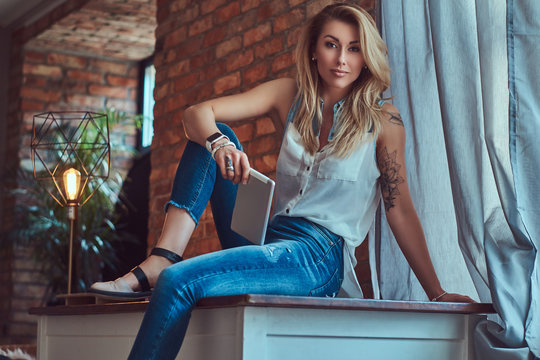 Fashionable blonde female blogger holds a tablet while sitting on a table against a brick wall in a studio with a loft interior.
