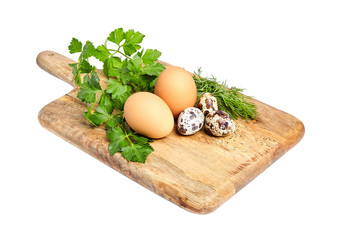 Chicken and spotted quail eggs on wooden cutting board. Green parsley and dill. White background