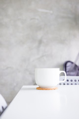 White cup on white table against light gray concrete wall. Minimalism concept. Layout, copyspace for text