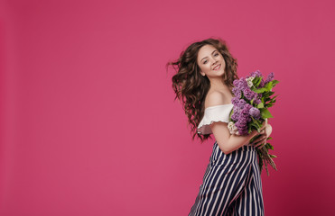 Obraz na płótnie Canvas Young beautiful girl with long wavy hair with a bouquet of purple flowers happy hurrying on a date. On a pink isolated background with space for text