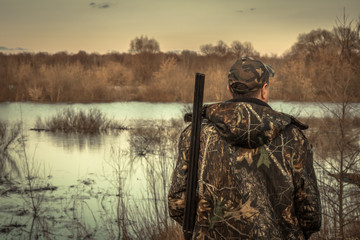 Hunter man shotgun camouflage exploring flood river hunting season rear view sunset