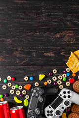 black and white modern gamepads on a dark wooden background among sweets, biscuits and cans of soda.