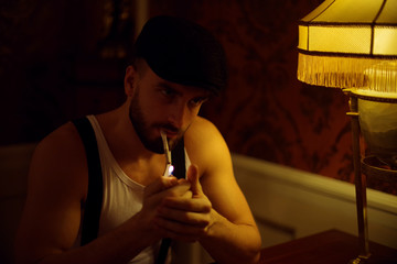 A handsome muscular man with a beard and a cap. Sitting in a white t-shirt in the Suite of an expensive hotel. Old school style. Gangster in English style. Lights a cigarette