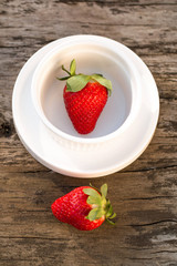 Close up, large, red, ripe strawberries. One is in a white bowl, the other is on a wooden table.
