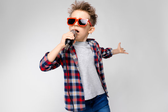 A handsome boy in a plaid shirt, gray shirt and jeans stands on a gray background. A boy wearing sunglasses. Red-haired boy sings into the microphone