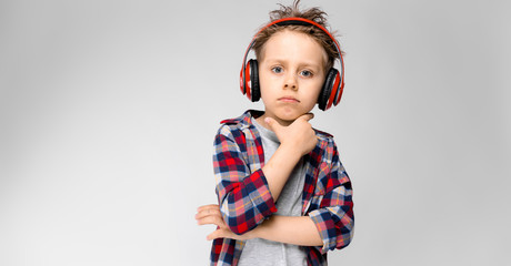 A handsome boy in a plaid shirt, gray shirt and jeans stands on a gray background. A boy in red headphones. The boy holds a hand to his chin.