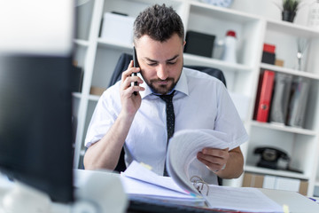 A man is sitting in the office, working with documents and talking on the phone.