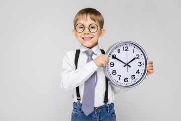 A charming boy in a white shirt, suspenders, a tie and light jeans stands on a gray background. The...