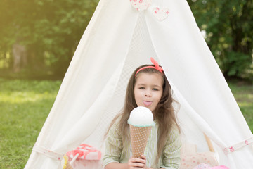Fototapeta na wymiar Little girl playing in a tent, children's house wigwam in park. Happy mothers day. Child with big donuts and ice cream and straw hat Summer, happy childhood concept. Cute emotions girl.