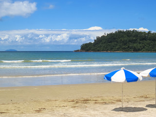 View of beach on sunny day with umbrella and island and clouds in the background.