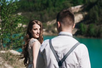 Fototapeta premium The bride in a beautiful dress holding hands with the groom in a light suit against the blue sky and blue water. Wedding couple standing on a sandy hill in the open air. A romantic love story.