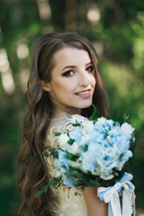 Naklejka premium Beautiful brunette bride with curls, makeup and stylish dress. Portrait on the background of green trees. Summer wedding day.