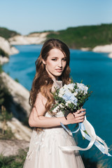 Beautiful brunette bride in white wedding dress with big long white train and with wedding bouquet stand on the edge of a cliff and a blue lake.  Summer vacation concept.