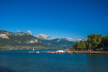 Fototapeta na wymiar View of Annecy lake, with island, vegetation, paddle boats, mountains and blue sky in background. Located in the department of Haute-Savoie, Auvergne-Rhone-Alpes region, southeastern France.