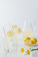 close up view of lemonade in jug and glasses with straws on white wooden tabletop on grey background