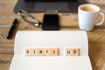 Closeup on notebook over wood table background, focus on wooden blocks with letters making Start Up word