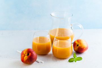 peach juice in glasses and a decanter and fresh peaches with leaves on a table. blue background. copy space