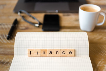 Closeup on notebook over wood table background, focus on wooden blocks with letters making Finance word