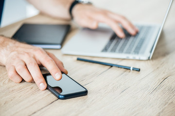 Fototapeta premium cropped shot of person holding smartphone and using laptop at wooden table