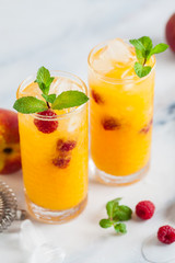two refreshing drinks from peach juice and raspberries with fresh mint in tall glasses on a marble table with a shaker and bar accessories.