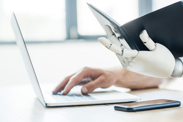 close-up view of robotic arm holding notebook and human hand using laptop at workplace