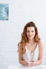 Obraz premium attractive young woman with cup of coffee in front of white brick wall looking at camera