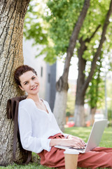 happy young freelancer working with laptop while leaning back on tree trunk in park and looking at camera