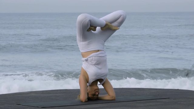 Medium shot of flexible mature woman doing handstand splits and lotus pose on coastline