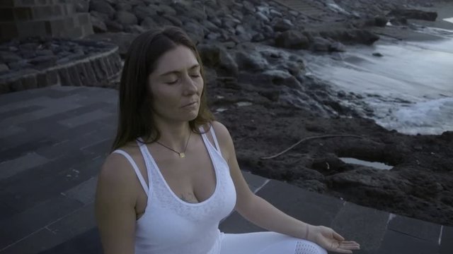PAN with zoom out of mindful woman in white sitting in lotus pose on stone dock near ocean and meditating in morning