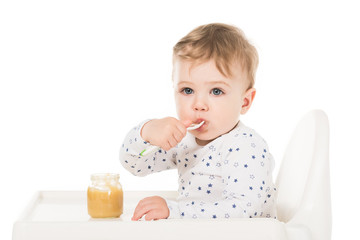  adorable baby boy eating puree from jar and sitting in highchair isolated on white background