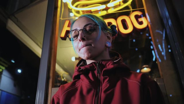 Hipster girl with blue dyed hair. Woman with piercing in nose, lenses, ears tunnels and unusual hairstyle stands at night illuminated city near neon showcase with hot dog