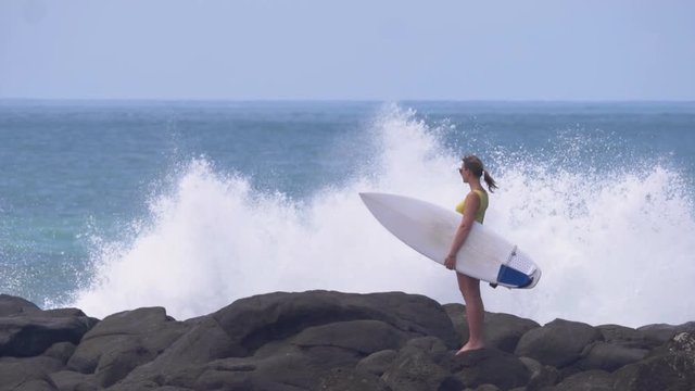 SLOW MOTION, COPY SPACE: Spectacular shot of water splashing while surfer girl stands still on a beautiful black beach. Powerful ocean waves hit the rocky shore. Female tourist looking into distance.