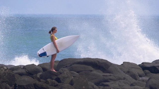 SLOW MOTION, COPY SPACE: Young surfer woman looks around the breathtaking sunny black beach. Splashing waves hit the coast on a beautiful summer day in New Zealand. Surfer waiting for good waves.