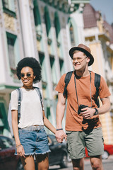 smiling multicultural couple of travelers with camera and backpacks holding hands of each other