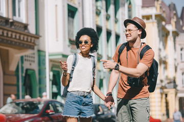 smiling interracial couple of travelers pointing by fingers to each other