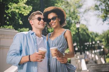 happy multiethnic couple in sunglasses with coffee cups standing on stairs in park