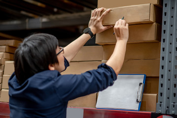 Young Asian man doing stocktaking of product in cardboard box on shelves in warehouse by using clipboard and pen. physical inventory count concept