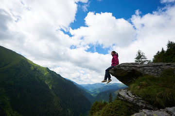 Silhouette of young female tourist sitting on the rock, dangling her legs in the abyss against the backdrop of green mountains with forests and clouds above them through which the blue sky is visible.
