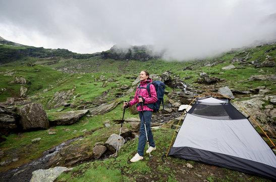 Smiling female tourist with backpack and walking sticks posing near tent on green grassy rocky meadow with beautiful mountains covered with fog in background. Romania Transfagarashan road.