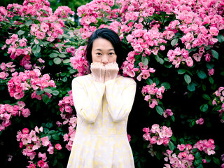  Outdoor portrait of beautiful young Chinese woman in yellow dress smiling among pink rose flowers wall in spring garden.
