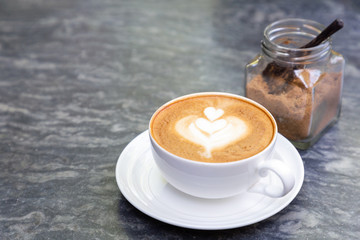 Cup of hot coffee with heart pattern in white cup and brown sugar on stone table background.
