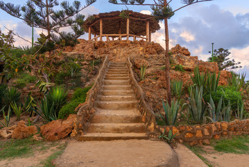Natural stone stairway with green bushes on both sides leading to wooden pergola with partly cloudy...