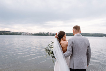 An amazing couple on their wedding day near the lake hug and enjoy each other. The bride and groom with a bouquet are happy together