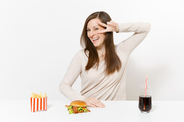Obraz na płótnie Canvas Happy woman shows victory sign at table with burger, french fries, cola in glass bottle isolated on white background. Proper nutrition or American classic fast food. Advertisement area with copy space