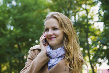 Beautiful girl with fair hair is talking on phone in green park. Young woman in scarf and beige coat. Cute autumn style photo.