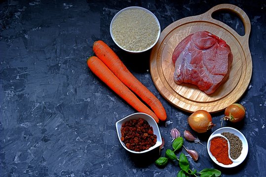 Ingredients for cooking pilaf on a dark gray concrete background. Meat on a wooden cutting board, carrots, rice, raisins, onion, garlic, spices.