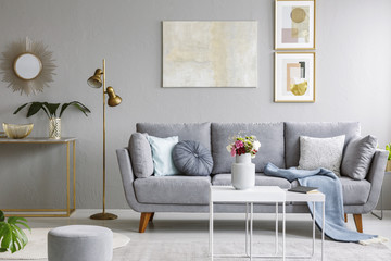 Gold mirror above shelf with plant in grey living room interior with sofa and flowers on table....