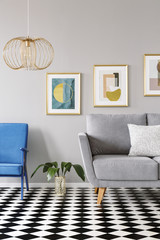 Gold lamp above blue armchair in checkered flat interior with grey settee and painting. Real photo
